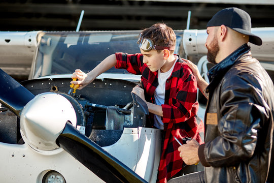 Aviation Male Engineer Giving Istructions To His Little Student Boy How To Execute Mechanic Fixing Of Disassembled Airplane Engine Block Of Light Single-engine Plane.