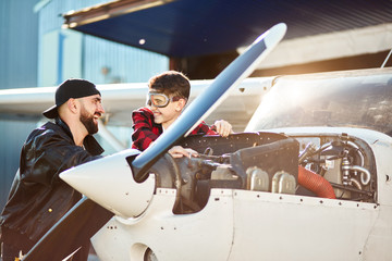 young handsome aircraft engineer and his little brother standing near single-engine propeller plane with opened motor cabinet, telling funny stories and laughing, enjoying time together.