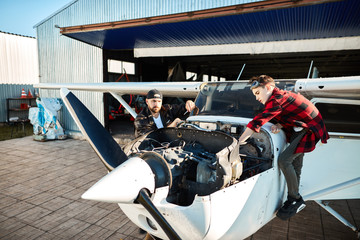 view of small white propeller air jet standing outside hangar building, male pilot with screwdriver stands besides, checking engine and his son helping him with inspecting airplane's systems.