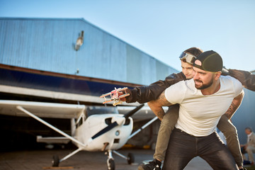 cheerful cute boy in large father's pilot jacket, sitting on dad's back, playing with toy plane, light private propeller plane and hangar on the background. Family and aviation concept. © alfa27