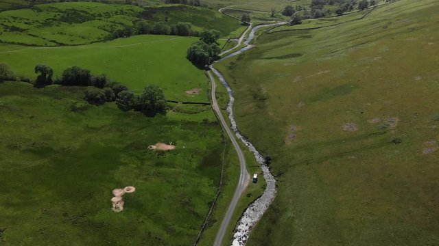 A high-altitude aerial shot looking down into the Barbon Beck Valley, Carnforth, Lancashire, England. This stunning countryside features a stream running alongside a country lane.