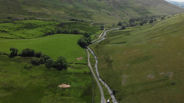 A high-altitude aerial shot looking down into the Barbon Beck Valley, Carnforth, Lancashire, England. This stunning countryside features a stream running alongside a country lane.