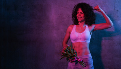 Portrait of a female athlete in a tracksuit standing over a concrete wall holding a bouquet of tulips, isolated over neon lights
