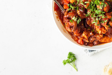 Meat stew with red beans, bell pepper and onion in tomato sauce in a white plate over light grey slate or stone background. 