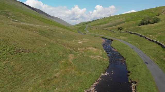 Flying over Barbon Beck Valley, Carnforth, Lancashire, England. This stunning countryside features a stream running alongside a small country lane where two cars are travelling into shot.