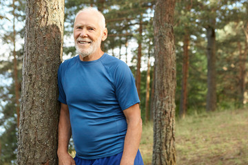 Naklejka na ściany i meble People, age, retirement, fitness and sports. Summertime image of positive happy seventy year old male pensioner catching breath during running exercise in pine forest, looking at camera with smile