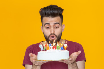 Close-up young handsome man blows off a candle from a burning cake posing for a yellow background. Holiday concept.