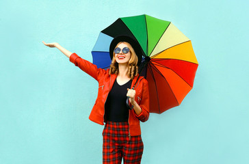 Obraz na płótnie Canvas Happy smiling young woman holding colorful umbrella in hands, wearing red jacket, black hat on blue wall background