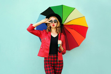 Stylish young woman with colorful umbrella blowing red lips sending sweet air kiss, wearing red...