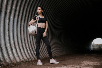 Sports woman posing in fashion sportswear on tunnel urban gray background. Fitness model working out outdoor. Beautiful slim girl in trendy black leggings, top and white backpack.