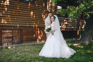 Fototapeta premium Beautiful newlyweds hugging and smiling against the background of a wooden house in the park with trees. Wedding portrait of a stylish groom in a beige suit and a cute bride in a dress with a bouquet.