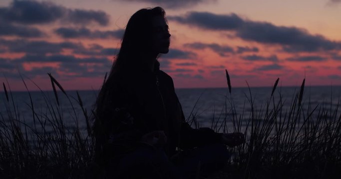 Black silhouette of meditating woman sitting by sea alone in violet light of dawn dusk. Unrecognizable young woman relaxing on ocean shore at summer vacation travel handheld device slow motion