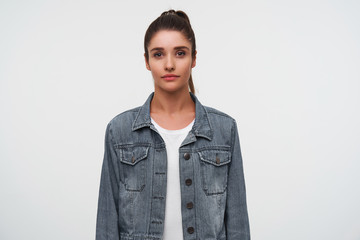 Portrait of young brunette lady wears in white t-shirt and denim jackets, looks at the camera with calm expression, stands over white background.