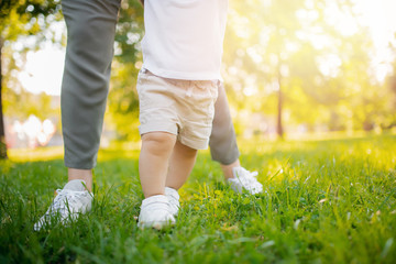 Child baby takes first step with help of mom in park. Concept parents teach walking