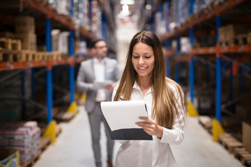 Portrait of gorgeous brunette woman manager controlling business in warehouse logistic center. Well dressed successful woman checking distribution while CEO walking behind her.