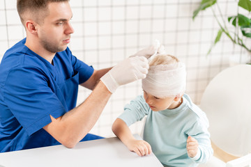 Obraz premium Doctor in blue uniform touching a boy head with trauma in his head and elastic bandaged around his head.
