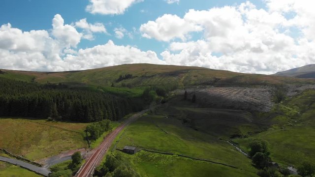 An aerial static shot looking at the Dent railway and the surrounding Cumbrian countryside. The scenery is green and sunny filmed in June. There are clouds passing over the landscape.