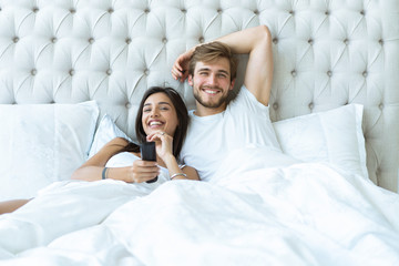 happy couple with remote lying in bed at home and watching tv.