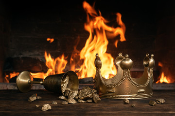 Golden crown and a goblet full of gold on the king table over burning fire background.