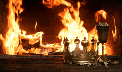 Golden crown and a goblet full of gold on the king table over burning fire background.