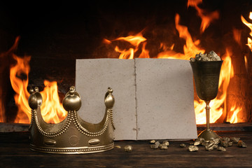Open blank page book, golden crown and a goblet full of gold on the king table over burning fire...