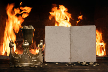 Open blank page book, golden crown and a goblet full of gold on the king table over burning fire...
