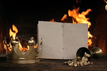 Open blank page book, golden crown and a goblet full of gold on the king table over burning fire...