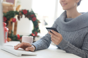 Close-up of satisfied young woman in gray sweater sitting at table and typing credit card details while paying for online purchase