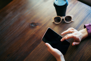 Overview of female hands, scrolling the screen of mobile phone. A cup of coffee and glasses beside.