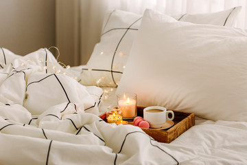 Wooden tray of coffee and candles on bed. White bedding sheets with striped blanket and pillow. Breakfast in bed. Hygge concept.