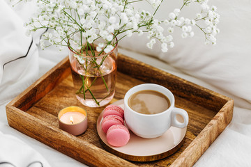 Wooden tray of coffee and candles with flowers on bed. White bedding sheets with striped blanket and pillow. Breakfast in bed. Hygge concept.