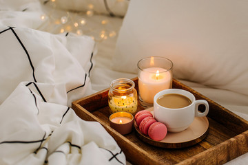 Wooden tray of coffee and candles on bed. White bedding sheets with striped blanket and pillow. Breakfast in bed. Hygge concept.