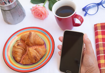 Coffee time for an elderly lady who enjoys the morning outside on the terrace. Cup of coffee, biscuits and a rose to brighten up the day. Positive moment. Looking at mobile phone