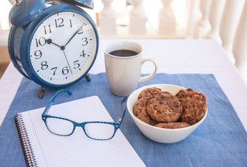 Breakfast or break outdoor on the balcony with coffe cup and chocolate biscuits.Big blue metal alarm clock. Notebook and eyeglasses