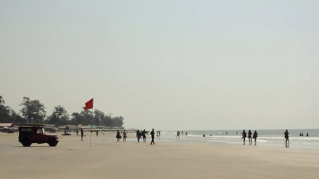 people tourists walk along the sandy beach and bathe in the sea waves and a lifeguard car near the red flag