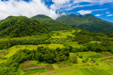 Aerial drone view of clouds passing over lush greenfarmland with mountains and volcanos in the background (Camiguin, Philippines)