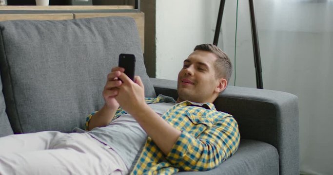 Handsome young man spends his free time with his smartphone, turns his phone from a vertical to a horizontal position, smiling, lying on a grey couch at home. Slow motion, 4K. Shot on RED camera.