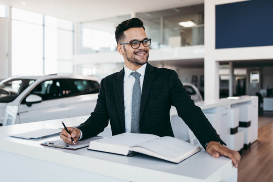 Good Looking, Cheerful And Friendly Salesman Poses In A Car Salon Or Showroom.