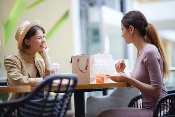 Happy fashionable young ladies sitting at round table on food court and discussing sales while...
