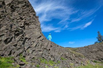 Fototapeta na wymiar A woman in a blue jacket stands on the stone mountain of the Hjalparfoss waterfall in Iceland in summer on a sunny day against a blue sky