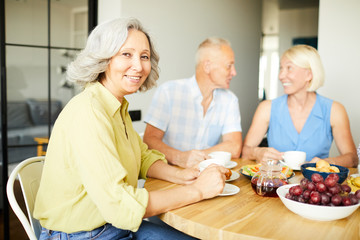 Portrait of elegant mature woman sitting at dinner table and smiling at camera while enjoying lunch with friends and family, copy space