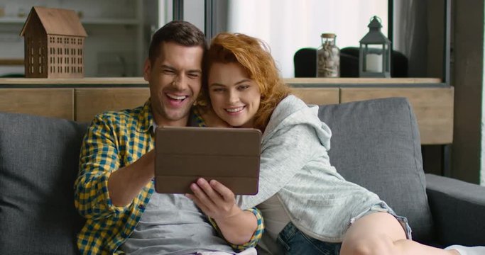 Happy young man and woman are watching photos on a tablet, smiling, recolecting funny moments from their life, having nice time together at home, sitting on a grey couch. 4K, shot on RED camera.