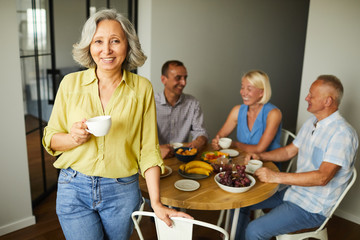 Portrait of beautiful senior woman smiling at camera posing at home with friends in background, copy space