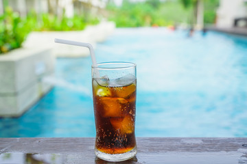 cola with ice in glass and white straw on wood beside swimming pool