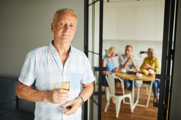 Obraz premium Waist up portrait of handsome mature man holding champagne flute looking at camera posing at home with friends in background, copy space