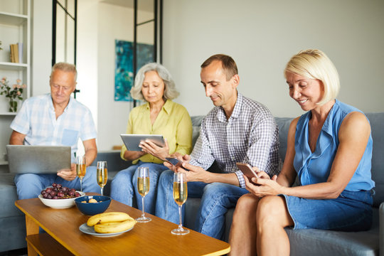 Group of contemporary senior people using various devices sitting on sofa at home, copy space