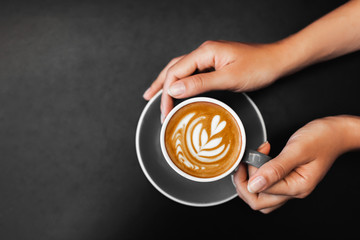 Cup of fresh cappuccino in woman hands on dark black metal table background. Empty place for text....
