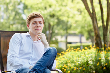 Young attractive man in white shirt sitting on brown chair in the park on green background - 279737711