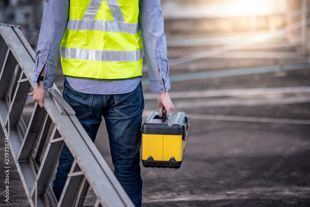 Wall mural Maintenance worker man with safety helmet and green vest carrying aluminium step ladder and tool box at construction site. Civil engineering, Architecture builder and building service concepts