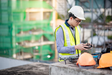 Asian engineer man working with drone, laptop and working tools at construction site. Male worker using unmanned aerial vehicle (UAV) for land and building site survey in civil engineering project.